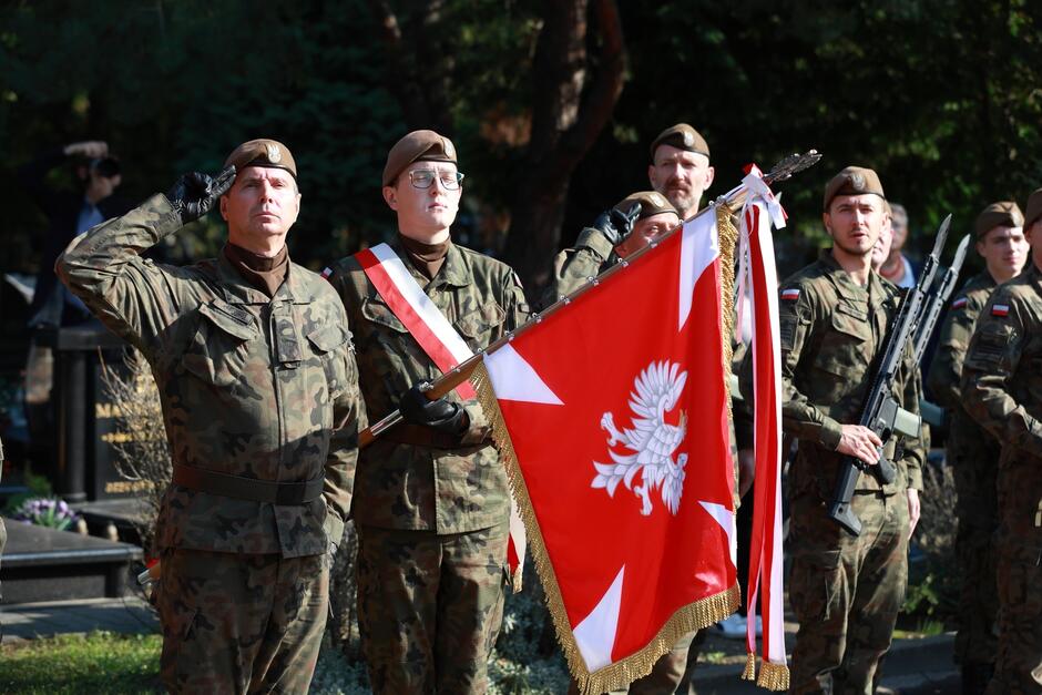 Na zdjęciu widoczna jest grupa żołnierzy w mundurach wojskowych, uczestniczących w oficjalnej uroczystości patriotycznej. Na pierwszym planie stoi żołnierz w pozycji salutującej, obok niego inny trzyma sztandar z orłem białym na czerwonym tle, symbolizujący narodowe barwy Polski. Sztandar jest ozdobiony złotymi frędzlami, co podkreśla jego oficjalny charakter. W tle znajdują się inni żołnierze w umundurowaniu, również stojący w pozycji honorowej. Widoczne są także elementy uzbrojenia, co świadczy o reprezentacyjnym charakterze tego oddziału. Cała scena ma podniosły i uroczysty charakter, sugerując, że jest to ceremonia upamiętniająca ważne wydarzenie historyczne lub narodowe święto.