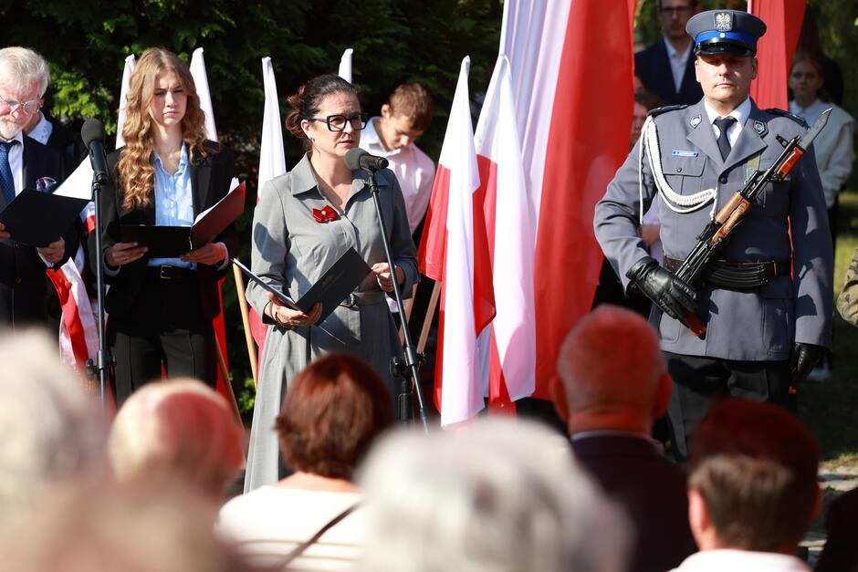 Na zdjęciu widoczna jest uroczystość patriotyczna, podczas której przemawia kobieta stojąca przy mikrofonie. Ubrana jest w szary strój, a na piersi ma przypięty czerwony symbol, co sugeruje, że może to być związane z obchodami historycznego wydarzenia lub święta narodowego. W dłoni trzyma czarną teczkę z dokumentami, z której prawdopodobnie czyta przemówienie. Po lewej stronie kobiety stoi młoda kobieta i starszy mężczyzna, oboje również trzymają dokumenty i są ubrani formalnie, co podkreśla oficjalny charakter wydarzenia. Po prawej stronie widoczny jest mężczyzna w mundurze wojskowym lub policyjnym, trzymający broń. W tle widać biało-czerwone flagi Polski, co wskazuje na patriotyczny wydźwięk uroczystości. Przed sceną zgromadzeni są ludzie, odwróceni tyłem do obiektywu, co sugeruje, że jest to ceremonia publiczna z udziałem widowni. Całość wskazuje na podniosłą i uroczystą atmosferę, prawdopodobnie związaną z uczczeniem ważnego wydarzenia historycznego.