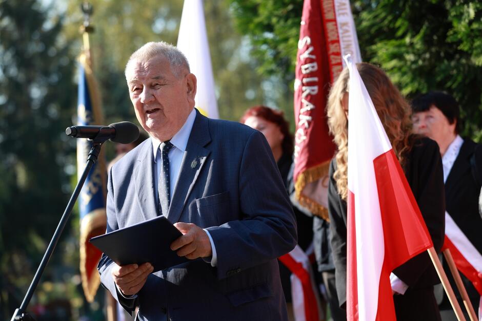 Na zdjęciu widoczny jest starszy mężczyzna w garniturze, stojący przy mikrofonie. Wygląda na to, że przemawia podczas uroczystości patriotycznej lub upamiętniającej. W dłoni trzyma ciemną teczkę z dokumentami, z której prawdopodobnie odczytuje swoje przemówienie. W tle widoczne są biało-czerwone flagi oraz sztandary, w tym jeden z napisem „Sybiracy,” co sugeruje, że wydarzenie jest związane z upamiętnieniem losów Polaków zesłanych na Syberię. Mężczyzna ma poważny, skupiony wyraz twarzy, co podkreśla podniosły charakter chwili. W otoczeniu widać również inne osoby w strojach formalnych, które dodają uroczystości oficjalnego charakteru.