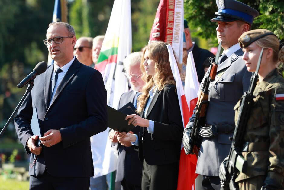 Na zdjęciu widoczna jest uroczystość patriotyczna. Na pierwszym planie stoi mężczyzna w garniturze przemawiający do mikrofonu, trzymający w dłoni teczkę z dokumentami. Jego wyraz twarzy jest poważny, co sugeruje, że przemawia w ważnej sprawie, prawdopodobnie związanej z upamiętnieniem wydarzeń historycznych. W tle widoczne są biało-czerwone flagi Polski oraz sztandary, w tym z napisem „Sybiracy,” co wskazuje na uroczystość związaną z pamięcią o losach Polaków zesłanych na Syberię. Obok niego stoi młoda kobieta i starszy mężczyzna, oboje w oficjalnych strojach, również z dokumentami w rękach. Po prawej stronie zdjęcia widać funkcjonariusza w mundurze policyjnym z bronią na ramieniu oraz kobietę w mundurze wojskowym, również z bronią. Obecność umundurowanych osób podkreśla uroczysty i oficjalny charakter wydarzenia. Cała scena odbywa się na świeżym powietrzu, prawdopodobnie w miejscu pamięci.