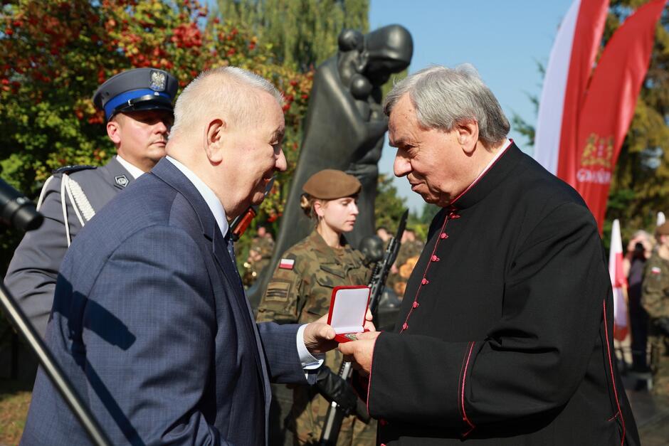 Na zdjęciu widoczna jest oficjalna uroczystość, podczas której starszy mężczyzna w garniturze wręcza medal księdzu w czarnej sutannie. Wydarzenie odbywa się na świeżym powietrzu, w otoczeniu innych uczestników. Mężczyźni stoją naprzeciw siebie, patrząc sobie w oczy, co sugeruje szacunek i powagę chwili. W tle widoczny jest pomnik oraz stojąca w pozycji honorowej kobieta w mundurze wojskowym z bronią na ramieniu. Obok nich stoi funkcjonariusz w mundurze policyjnym, również w postawie oficjalnej. Czerwone flagi i zielona roślinność tworzą tło, podkreślając uroczysty charakter wydarzenia, które najprawdopodobniej upamiętnia ważne historyczne wydarzenie lub osobę.