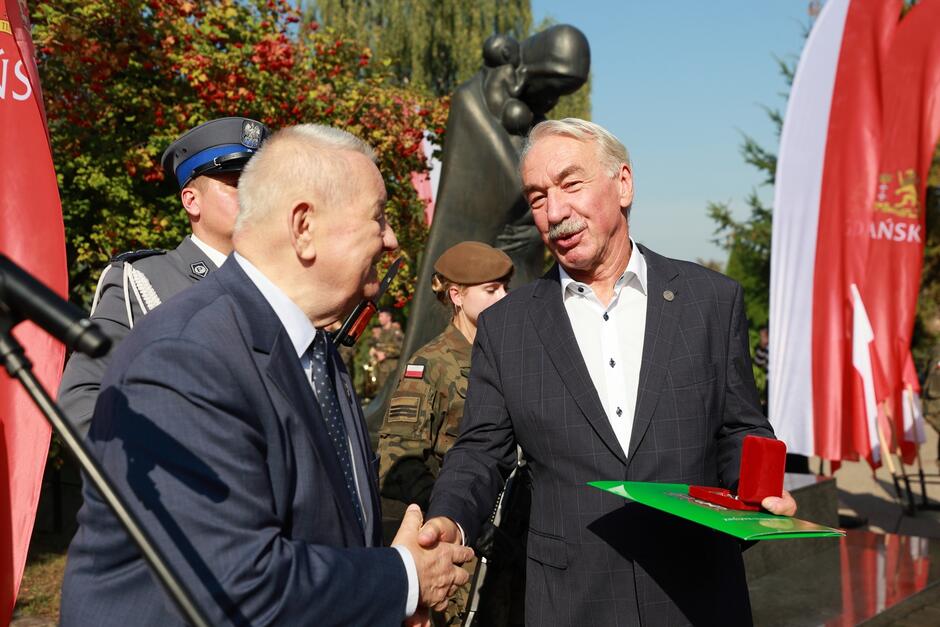 Na zdjęciu widać moment uroczystego wręczenia odznaczenia. Dwóch mężczyzn, ubranych w eleganckie garnitury, podaje sobie ręce w geście gratulacji. Mężczyzna po lewej stronie, starszy, trzyma w dłoni medal, który wręcza drugiemu mężczyźnie po prawej stronie. Mężczyzna po prawej stronie trzyma zieloną teczkę oraz pudełko z odznaczeniem, wyrażając zadowolenie i wdzięczność. W tle widoczna jest osoba w mundurze wojskowym, a także pomnik upamiętniający, co sugeruje, że ceremonia ma miejsce w miejscu pamięci narodowej. Po lewej stronie zdjęcia można dostrzec fragment mikrofonu, co wskazuje, że wydarzenie może być rejestrowane lub jest częścią większej uroczystości. Otoczenie z czerwonymi flagami oraz obecność funkcjonariusza w mundurze policyjnym podkreślają oficjalny i patriotyczny charakter tego wydarzenia.