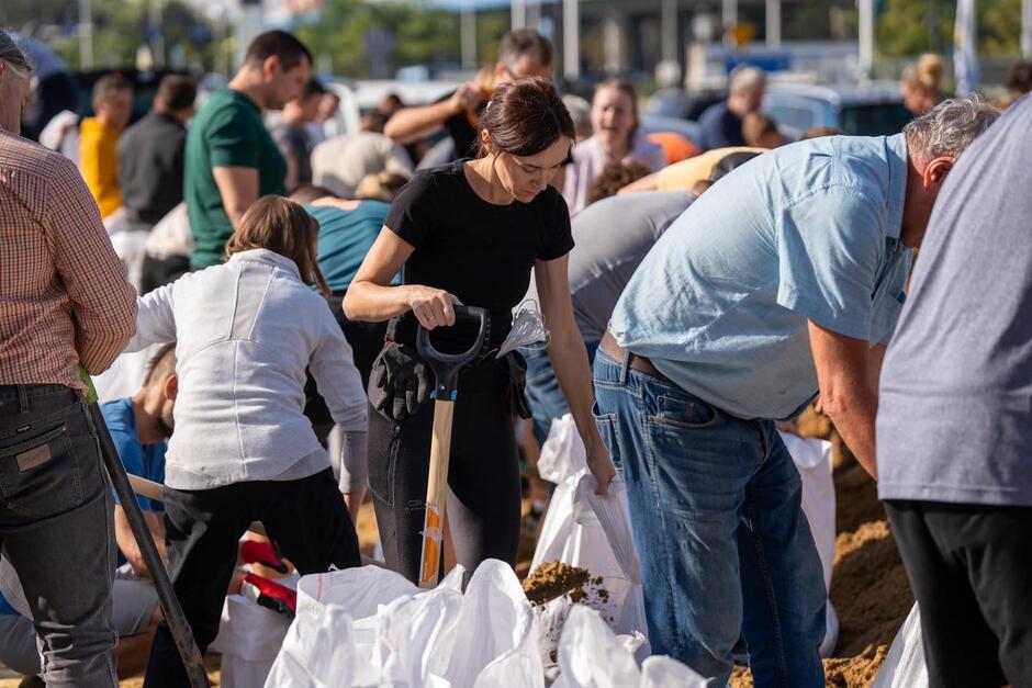 Na zdjęciu widać grupę ludzi pracujących razem nad napełnianiem worków z piaskiem, prawdopodobnie w ramach akcji pomocowej lub zabezpieczania przed powodzią. W centrum kadru kobieta w czarnej koszulce trzyma łopatę, koncentrując się na swoim zadaniu.