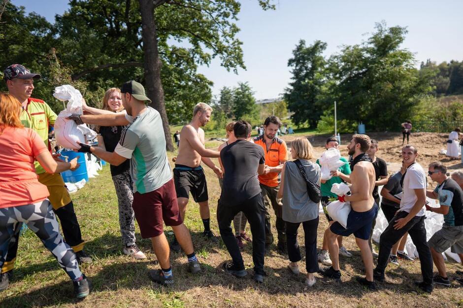 Grupa młodych ludzi ustawiona jest w rzędzie, przekazują sobie worki z piaskiem.