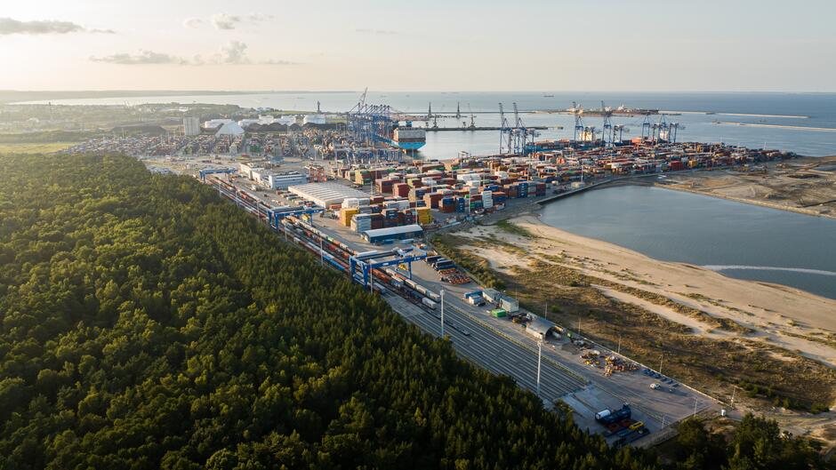 Zdjęcie przedstawia duży obszar portowy, prawdopodobnie terminal kontenerowy, położony nad zbiornikiem wodnym. Widać na nim liczne dźwigi oraz stosy kolorowych kontenerów, co sugeruje, że port jest bardzo aktywny. Wzdłuż portu przebiega linia kolejowa oraz droga, co wskazuje na zintegrowany węzeł transportowy. Po jednej stronie zdjęcia rozciąga się gęsty las, który graniczy z infrastrukturą portu, tworząc kontrast między naturalnym krajobrazem a przemysłowym obszarem.