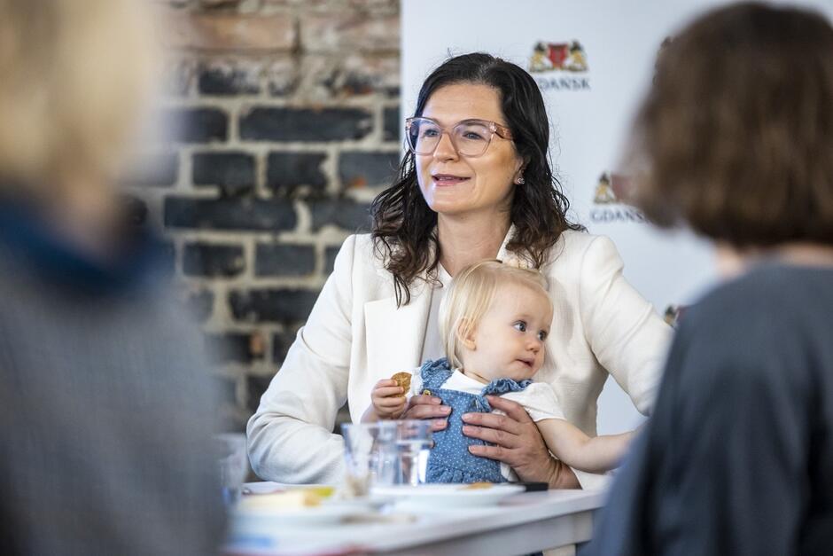 Na zdjęciu znajduje się kobieta o ciemnych, falowanych włosach, nosząca okulary i ubrana w elegancką, jasną marynarkę. Kobieta trzyma na kolanach małe dziecko ubrane w niebieski kombinezon, które również było widoczne na poprzednim zdjęciu. Dziecko jest przytulone do kobiety i patrzy w bok, trzymając w rączce przekąskę. W tle widoczna jest ceglana ściana oraz logo miasta Gdańsk, co sugeruje, że zdjęcie zostało zrobione podczas jakiegoś wydarzenia związanego z tym miastem. Na pierwszym planie widać rozmyte sylwetki innych osób. 