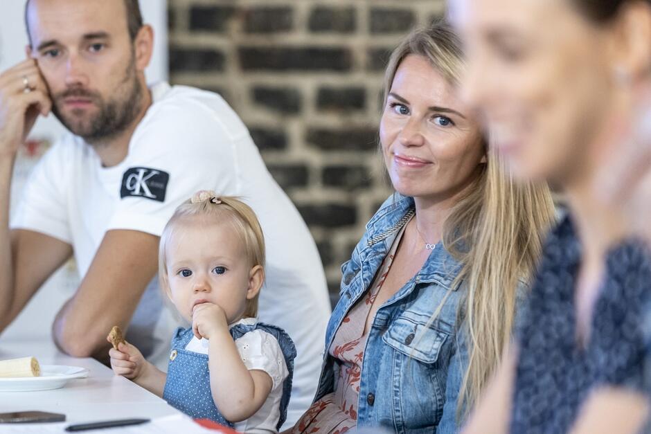 Na zdjęciu widać grupę osób siedzących przy stole. W centrum znajduje się kobieta z długimi, blond włosami, która patrzy w stronę obiektywu i lekko się uśmiecha. Obok niej siedzi małe dziecko z jasnymi włosami, ubrane w niebieski kombinezon, które je przekąskę i również patrzy w kierunku aparatu. W tle z lewej strony znajduje się mężczyzna w białej koszulce, wspierający głowę na ręce i patrzący przed siebie. Na pierwszym planie po prawej stronie widać sylwetkę innej osoby, która jest nieco rozmyta. W tle widać ceglaną ścianę.