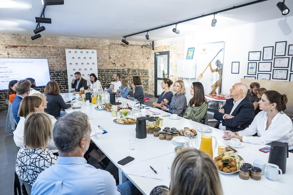 Na zdjęciu widać dużą grupę osób siedzących przy długim stole ustawionym w sali konferencyjnej. Na stole znajduje się różnorodne jedzenie, w tym talerze z przekąskami, słoiczki, dzbanki z napojami i filiżanki, sugerujące, że odbywa się tu spotkanie w formie śniadania prasowego lub konferencji.
W tle można dostrzec ceglaną ścianę oraz banery z logo miasta Gdańsk. Na ścianie widoczne są również ramki z rysunkami, co dodaje wnętrzu przytulnego charakteru. Wśród uczestników spotkania znajduje się kilka osób w formalnym stroju, a także kobieta z ciemnymi włosami i okularami, która trzyma małe dziecko. Na jednym z ekranów po lewej stronie widnieją napisy oraz dane, sugerujące, że jest to oficjalne wydarzenie lub prezentacja.