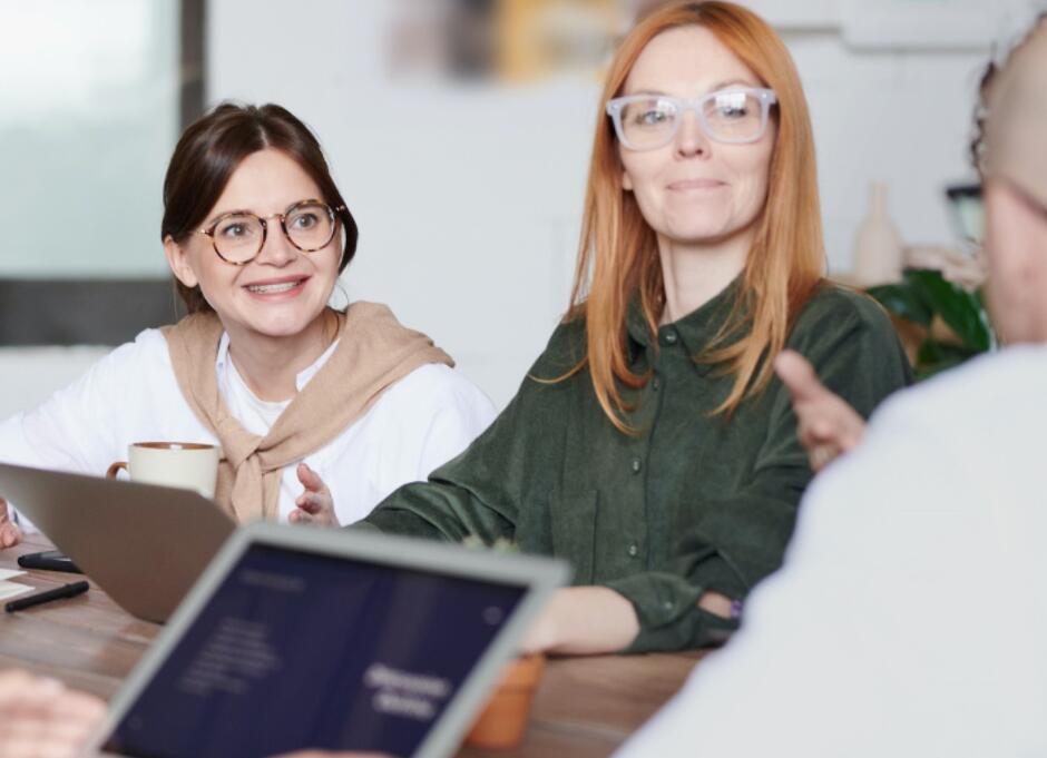 Na zdjęciu widoczne są dwie kobiety siedzące przy stole w jasnym, nowoczesnym pomieszczeniu. Kobieta po lewej stronie ma ciemne włosy spięte w kok, nosi okulary o okrągłych oprawkach i uśmiecha się. Ubrana jest w białą koszulę, na której ma zarzucony beżowy sweter. Na stole przed nią stoi laptop, co wskazuje, że może brać udział w rozmowie biznesowej lub dyskusji. Kobieta po prawej stronie ma długie, rude włosy i nosi okulary o przezroczystych oprawkach. Ubrana jest w ciemnozieloną koszulę. Patrzy w stronę osoby, która znajduje się poza kadrem, i ma lekko zamyślony wyraz twarzy. Na pierwszym planie, częściowo widoczny, znajduje się ekran laptopa osoby siedzącej naprzeciwko, co sugeruje, że zdjęcie zostało wykonane podczas spotkania lub rozmowy.