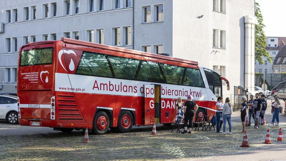 Na zdjęciu widać autobus do poboru krwi zaparkowany przed budynkiem. Autobus jest w kolorach czerwono-białych, z napisem "Ambulans do pobierania krwi" oraz nazwą "GDAŃSK", wskazującą na przynależność regionalną. Z boku autobusu znajduje się także logo Regionalnego Centrum Krwiodawstwa i Krwiolecznictwa w Gdańsku. Przed autobusem zgromadzili się ludzie – prawdopodobnie część z nich przygotowuje się do oddania krwi, a reszta pomaga w organizacji. Miejsce zdarzenia wydaje się być przestrzenią publiczną, prawdopodobnie w mieście, sądząc po otaczających budynkach i zaparkowanych samochodach. Cała scena sugeruje lokalną akcję krwiodawstwa.