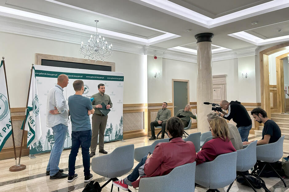 Konferencja prasowa w holu eleganckiego budynku z kolumnami. Mężczyzna mówi do mikrofonu, na fotelach siedzą ludzie i słuchają.