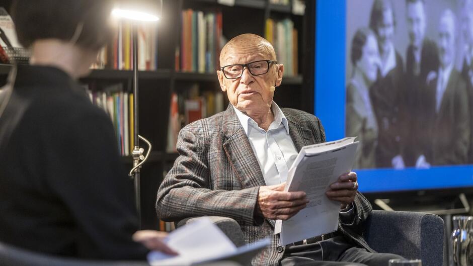 Na zdjęciu widzimy starszego mężczyznę w eleganckiej marynarce i okularach, siedzącego na fotelu, prawdopodobnie podczas wywiadu lub dyskusji w studiu lub bibliotece. Mężczyzna trzyma w rękach kilka kartek z dokumentami, co sugeruje, że jest zaangażowany w rozmowę na temat ważnych tematów, być może związanych z historią lub kulturą. Ma na sobie mikrofon przypięty do ucha, co wskazuje na to, że bierze udział w nagrywanym lub transmitowanym wydarzeniu. W tle widoczny jest regał z książkami oraz ekran, na którym wyświetlone jest czarno-białe zdjęcie grupy ludzi, co może wskazywać na kontekst historyczny omawianych tematów. Całość tworzy atmosferę intelektualnej debaty lub wykładu.