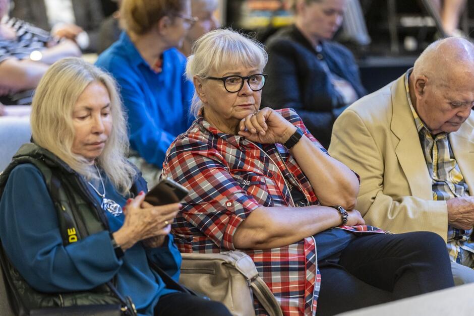 Na zdjęciu widzimy grupę starszych osób siedzących w rzędach, prawdopodobnie podczas wydarzenia, takiego jak wykład, spotkanie autorskie lub wywiad. Na pierwszym planie widoczna jest kobieta w koszuli w kratę, która patrzy skupiona przed siebie, wspierając rękę na brodzie. Obok niej siedzi inna kobieta, ubrana w ciemniejszą kurtkę, która trzyma w ręku telefon, oraz starszy mężczyzna w jasnej marynarce, który pochyla głowę i wygląda, jakby drzemnął. W tle widać inne osoby, również skupione na tym, co dzieje się przed nimi. Atmosfera zdjęcia jest spokojna, a uczestnicy wydają się być zaangażowani w obserwowane wydarzenie.