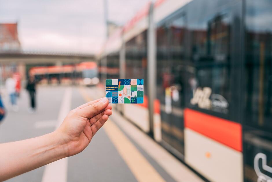 Na zdjęciu widzimy osobę trzymającą w ręce kolorową kartę z napisem Gdańska Karta Mieszkańca  na tle tramwaju miejskiego