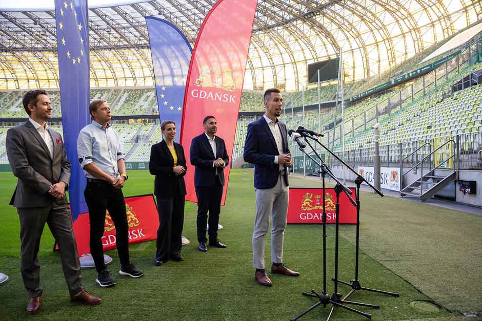 Mężczyzna mówi do mikrofonu na stadionie podczas konferencji prasowej. Za nim stoi trzech innych mężczyzn i kobieta.