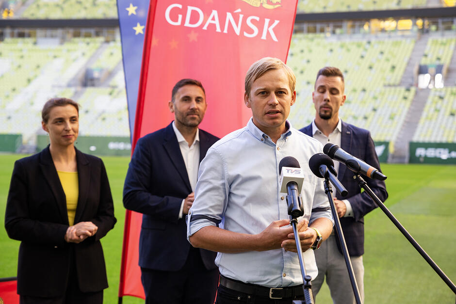 Mężczyzna mówi do mikrofonu podczas konferencji prasowej na stadionie. Za nim stoi dwóch innych mężczyzn i kobieta.