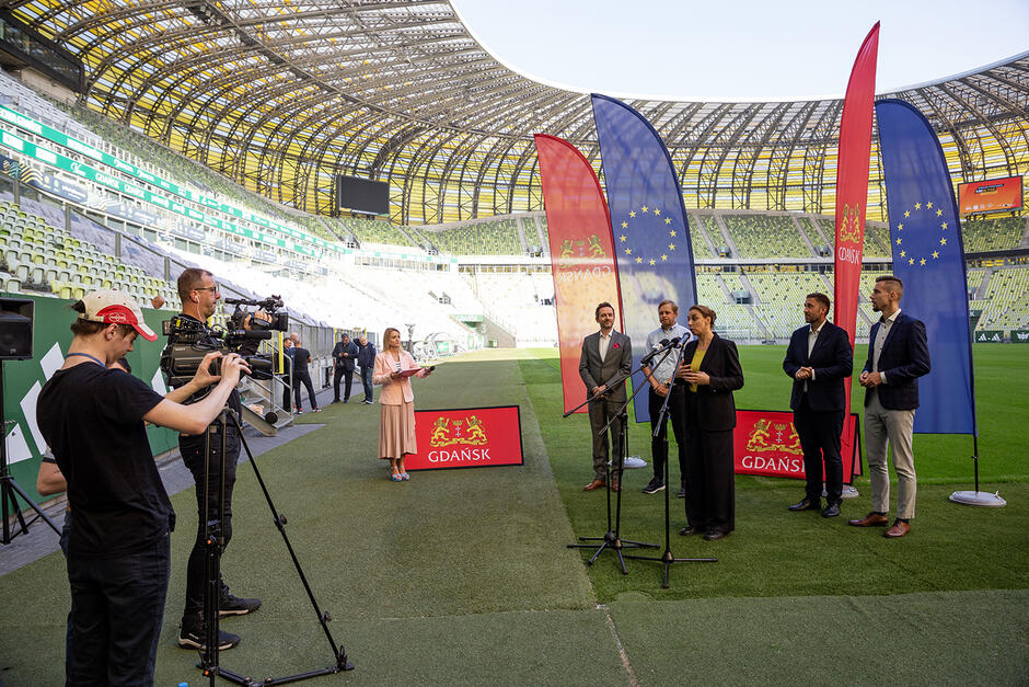 Konferencja prasowa na stadionie. Przed grupą dziennikarzy stoi czterech mężczyzn i kobieta, która mówi do kimrofonu.