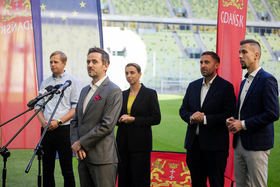 Czterech mężczyzn i kobieta stoją przed mikrofonami na stadionie podczas konferencji prasowej.