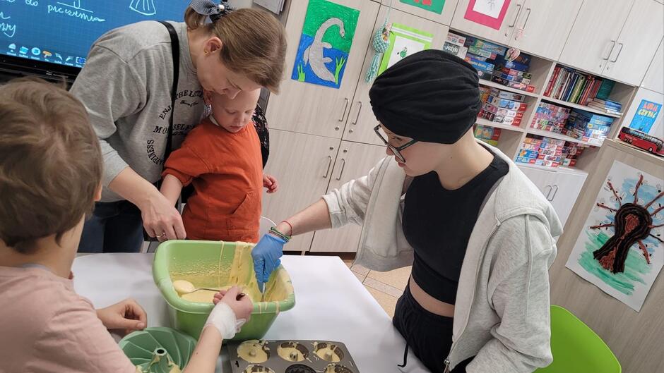 Na zdjęciu widzimy grupę osób w pomieszczeniu, prawdopodobnie klasie lub sali terapeutycznej, zajmujących się przygotowywaniem jedzenia, najpewniej wypieków. Na pierwszym planie widoczna jest miska z jasnym ciastem, które jest nakładane do formy. Jedna osoba, ubrana w czarny top i szarą bluzę z kapturem, nosi na głowie czarną chustę lub czapkę, a także ma założoną niebieską rękawiczkę ochronną i pomaga przy mieszaniu ciasta. Obok tej osoby stoi kobieta pochylająca się nad małym dzieckiem w pomarańczowej koszulce, które z zainteresowaniem obserwuje to, co się dzieje. Kobieta, która ma związane włosy w kitkę, ubrana jest w szarą bluzę z napisami i wspiera dziecko w czynności. W tle widać innego dziecka z tyłu, które jest skierowane tyłem do aparatu. 