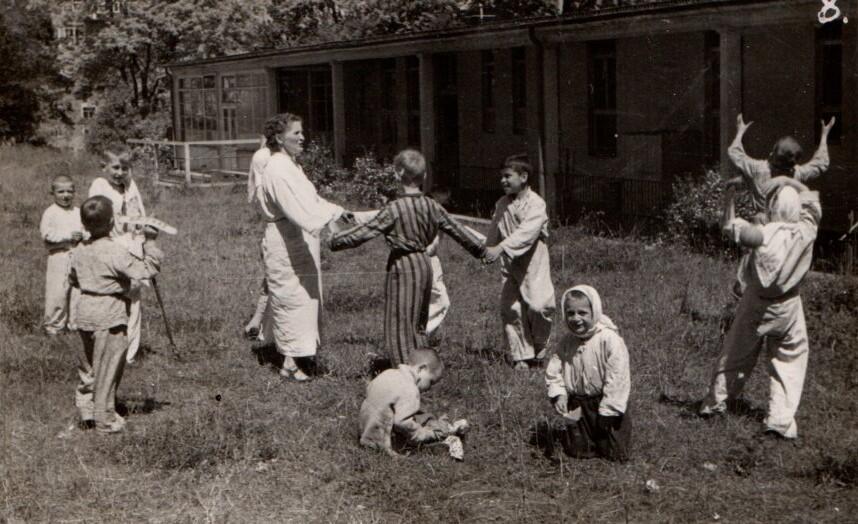 sanatorium lub ośrodku zdrowia. Dzieci są ubrane w piżamy i szlafroki, co sugeruje, że są pacjentami tej placówki. Większość z nich bierze udział w zabawie na trawie, trzymając się za ręce i tworząc krąg. Po lewej stronie dwoje dzieci stoi z kijkami, być może wspierając się nimi podczas chodzenia, co może wskazywać na rehabilitację. W centrum zdjęcia widoczna jest kobieta w białym fartuchu, która wydaje się nadzorować zabawę lub prowadzić aktywności z dziećmi. Na pierwszym planie, po prawej stronie, jedno z dzieci klęczy na trawie, patrząc w stronę aparatu, a obok siedzi kolejne, zajęte czymś na ziemi. W tle widoczny jest budynek, który wygląda jak pawilon sanatoryjny lub szpitalny, otoczony zielenią. Cała scena rozgrywa się na zewnątrz, w słoneczny dzień, co nadaje zdjęciu radosny i swobodny charakter, mimo że przedstawia dzieci najprawdopodobniej przebywające w ośrodku zdrowotnym.