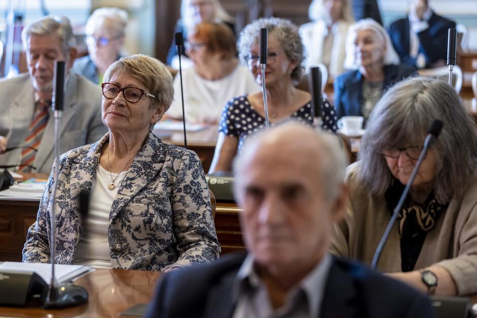 na zdjęciu widać kilku starszych seniorów, siedzą za stołami, na pierwszym planie starsza kobieta w eleganckiej marynarce i w okularach