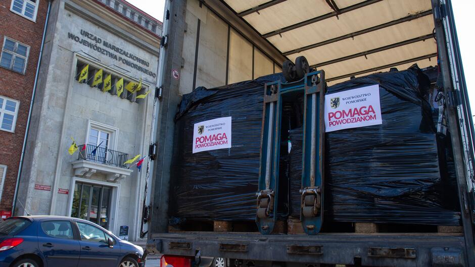 Ciężarówka wypełniona towarami, które widać przez szeroko otwarte drzwi pojazdu. Samochód stoi na ulicy przed zabytkowym budynkiem z napisem: Wojewódzki Urząd Marszałkowski 