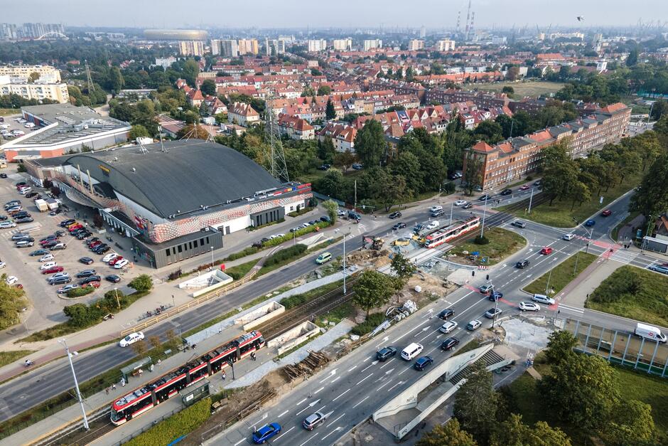 zdjęcie z drona, widać fragment dwóch jezdni, po lewej duży budynek centrum handlowego, w tle za nim gęsto zabudowane osiedle domów jednorodzinnych z czerwonymi dachami