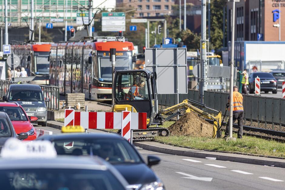 na zdjęciu sporo się dzieje, widać na pierwszym planie robotników wykonujących prace ziemne, jeden siedzi w małej koparce, drugi trzyma łopatę, za nimi widać jadący tramwaj biało czerwony, przed nimi w dolnej części zdjęcia widać fragmenty kilku samochodów jadących ulicą