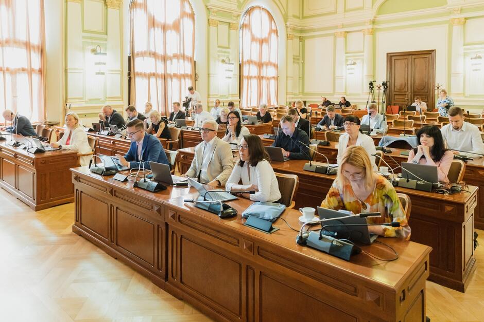 Sala rady miasta podczas obrad. Radni siedzą rzędami przy biurkach