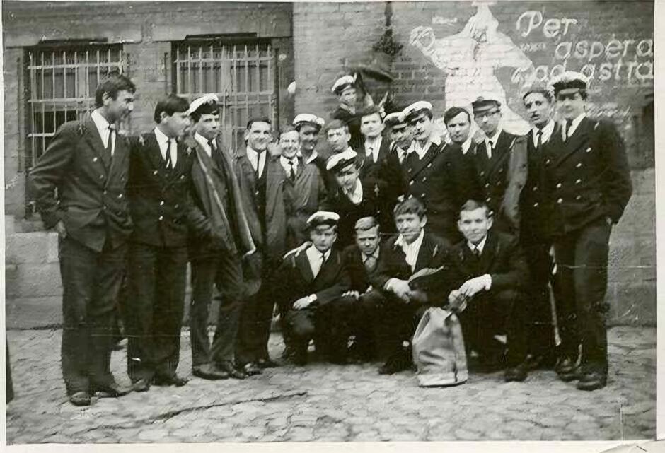 Zdjęcie przedstawia grupę maturzystów z klasy A z roku 1965, którzy uczęszczali do szkoły Conradinum. Uczniowie są ubrani w formalne mundury marynarskie, co sugeruje, że szkoła mogła mieć profil techniczno-morski lub wojskowy. Większość z nich ma na głowach charakterystyczne czapki marynarskie, a część trzyma je w rękach. Grupa stoi przed budynkiem szkoły, który ma ceglaną ścianę, na której widnieje namalowany napis łaciński  Per aspera ad astra , co oznacza  Przez trudy do gwiazd . To motto często stosowane w kontekście edukacyjnym, motywujące uczniów do przezwyciężania trudności w drodze do sukcesu.  Na zdjęciu panuje raczej nieformalna atmosfera, mimo oficjalnych mundurów – wielu uczniów uśmiecha się, a niektórzy zajmują różne pozycje, siedząc lub stojąc w luźnej grupie. Zdjęcie wydaje się być pamiątkowe, upamiętniające moment zakończenia szkoły i matury.