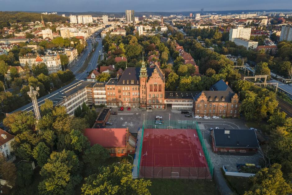 Zdjęcie przedstawia widok z lotu ptaka na kompleks szkolny Conradinum w Gdańsku. W centralnej części fotografii widać główny budynek szkoły – imponujący, ceglany gmach o klasycznej architekturze, z charakterystyczną wieżą zegarową. Po jego prawej stronie znajdują się inne budynki, również wykonane z czerwonej cegły, harmonizujące z głównym gmachem. Na pierwszym planie widoczne jest nowoczesne boisko sportowe z czerwonym nawierzchnią, przeznaczone do gry w piłkę nożną oraz inne sporty. Boisko jest otoczone siatką ochronną, a obok niego znajdują się dodatkowe budynki użytkowe. Otoczenie szkoły jest pełne zieleni, w tle widać gęsty las, a wokół rozciągają się budynki mieszkalne i infrastruktura miejska. Widoczna droga biegnąca po lewej stronie prowadzi w kierunku centrum miasta, a za nią widać dachy kamienic i nowoczesnych bloków. Na horyzoncie majaczy panorama miasta, z wysokimi budynkami, co podkreśla urbanistyczny charakter okolicy. Zdjęcie ukazuje połączenie tradycyjnej architektury szkolnej z nowoczesnymi udogodnieniami sportowymi, a także zieloną i przyjazną okolicę, w której znajduje się szkoła.