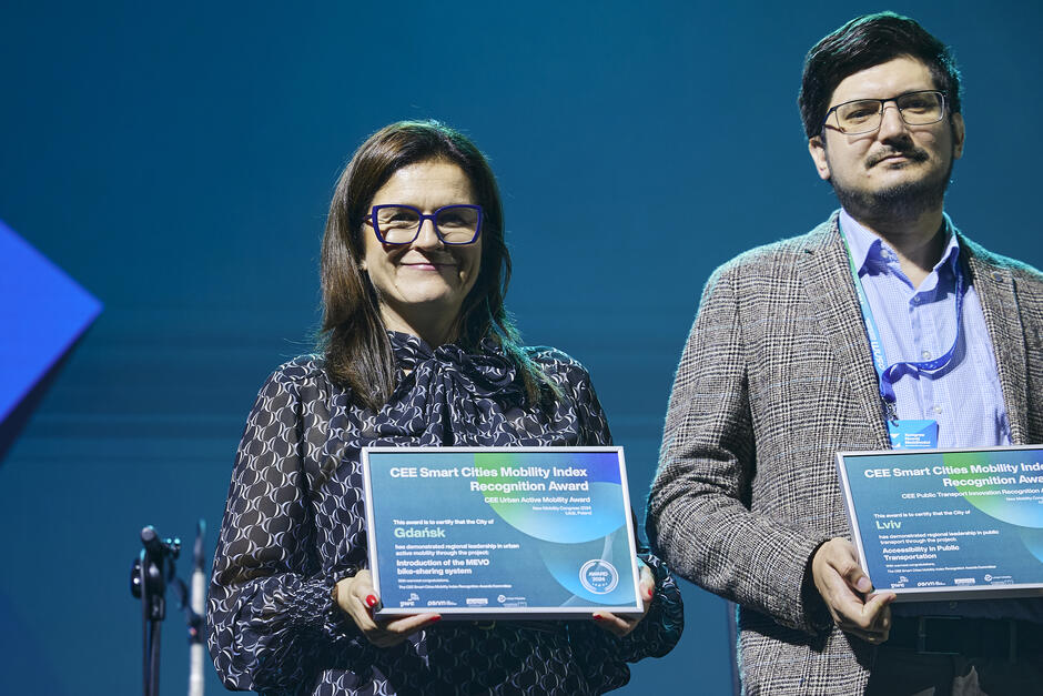 Na zdjęciu widoczne są dwie osoby, które trzymają certyfikaty za wyróżnienia w ramach CEE Smart Cities Mobility Index Recognition Award. Certyfikat dla miasta Gdańsk dotyczy wprowadzenia systemu rowerów miejskich MEVO, natomiast certyfikat dla miasta Lwów odnosi się do dostępności w transporcie publicznym.