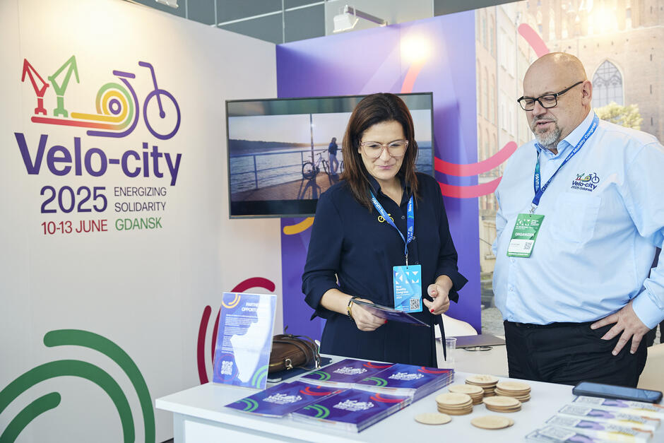 Na zdjęciu widzimy stoisko promujące konferencję Velo-city 2025, która odbędzie się w dniach 10-13 czerwca w Gdańsku. Dwie osoby w biznesowych strojach, wyposażone w identyfikatory, przeglądają materiały promocyjne na temat wydarzenia, które dotyczy mobilności rowerowej i zrównoważonego transportu.