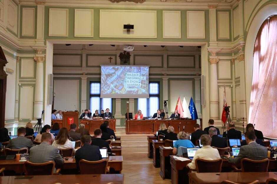 Zdjęcie przedstawia sesję rady miejskiej w eleganckiej sali obrad, w której uczestnicy siedzą przy stołach z komputerami, zwróceni w stronę prezydium. Na dużym ekranie widoczny jest slajd z napisem „Bieżące informacje o życiu Gdańska”, a za stołem prezydialnym znajdują się flagi Polski, Gdańska i Unii Europejskiej