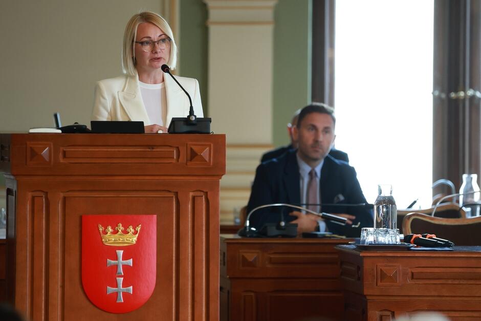 Na zdjęciu widzimy kobietę w białej marynarce, stojącą przy mównicy z herbem Gdańska, przemawiającą podczas oficjalnej sesji lub spotkania. W tle siedzi mężczyzna, uważnie słuchający, z rękami złożonymi na stole, na którym znajduje się szklana karafka i mikrofony