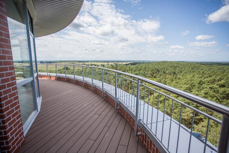 Zdjęcie przedstawia przestronny taras widokowy, znajdujący się na górze wieży ciśnień. Taras jest otoczony metalową balustradą, a podłoga wykonana jest z drewnopodobnych desek, co nadaje miejscu elegancki i nowoczesny wygląd. Po lewej stronie widać szklaną fasadę budynku, odbijającą widok na zewnątrz. Z tarasu roztacza się szeroki, panoramiczny widok na okolicę, który obejmuje rozległe lasy oraz pola, sięgające po horyzont. W pogodny dzień, z błękitnym niebem i rozproszonymi białymi chmurami, sceneria wygląda spokojnie i malowniczo. Widok ten sugeruje, że wieża ciśnień znajduje się w podmiejskim lub wiejskim otoczeniu, otoczonym przyrodą. To miejsce z pewnością oferuje wspaniałą przestrzeń do podziwiania krajobrazu z wysokości.