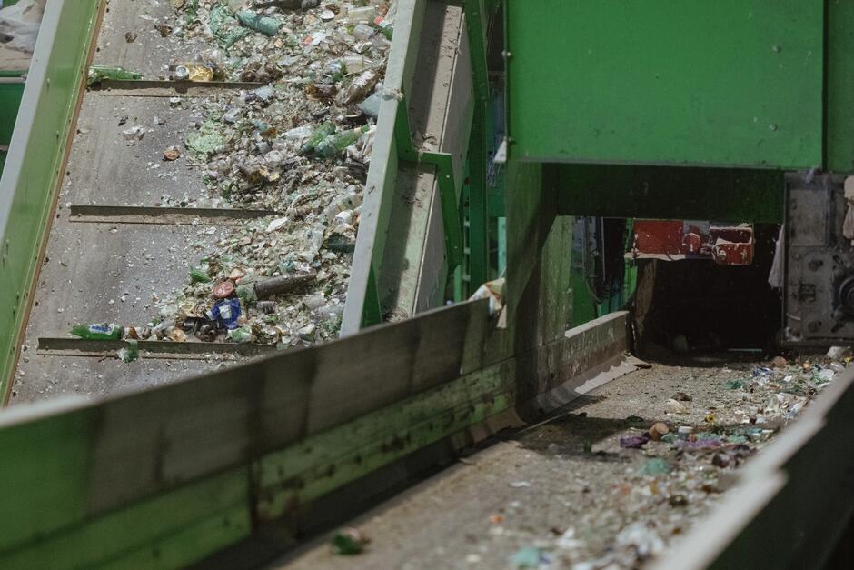 Na zdjęciu przedstawiono taśmociąg w zakładzie recyklingu szkła, na którym znajdują się rozbite butelki oraz inne odpady szklane. Taśma przesuwa materiał w stronę maszyny lub dalszej części procesu, co sugeruje, że jest to etap transportu szkła do dalszej obróbki lub sortowania.