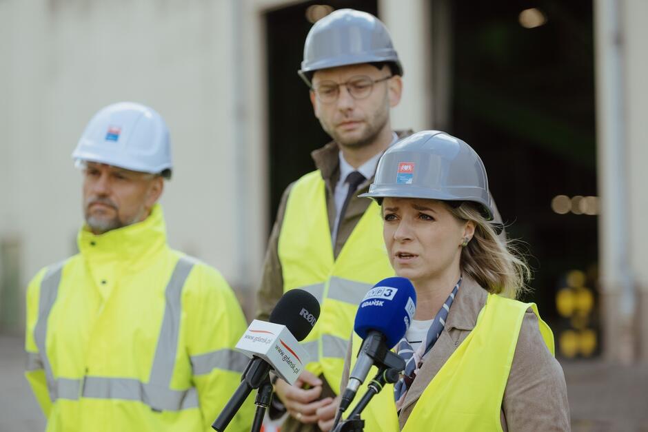 Na zdjęciu widzimy trzy osoby w kaskach ochronnych i kamizelkach odblaskowych, uczestniczące w konferencji prasowej. Kobieta na pierwszym planie wypowiada się do mikrofonów, podczas gdy dwóch mężczyzn stoi za nią, sugerując oficjalne wydarzenie związane z przemysłem lub recyklingiem.