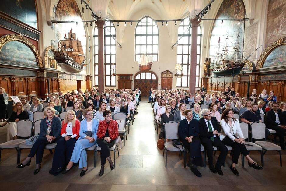 Na zdjęciu widzimy dużą grupę ludzi siedzących w zabytkowej sali, prawdopodobnie uczestników oficjalnej uroczystości, jak wręczenie nominacji czy nagród. Sala jest ozdobiona modelami statków oraz malowidłami, a uczestnicy siedzą w rzędach, zwróceni w stronę sceny lub mównicy.