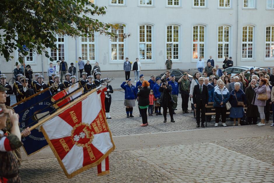 Na zdjęciu widać scenę uroczystości patriotycznej na świeżym powietrzu, w której bierze udział liczna grupa osób. Na pierwszym planie widoczne są sztandary, trzymane przez żołnierzy lub członków organizacji kombatanckiej. Jeden ze sztandarów ma napis „Armia Krajowa” oraz biało-czerwone barwy z orłem. Wokół sztandarów stoją osoby w mundurach, zarówno marynarze, jak i harcerze, którzy oddają honory poprzez salutowanie. W tle widać większą grupę uczestników, w tym ludzi w różnym wieku, którzy biorą udział w ceremonii. Niektórzy z nich stoją wyprostowani, inni wykonują gesty oddania honoru, a jeszcze inni fotografują lub obserwują przebieg uroczystości. Budynek w tle, z dużymi oknami, wygląda na oficjalny lub historyczny obiekt, co podkreśla formalny charakter wydarzenia. Całość sprawia wrażenie uroczystości upamiętniającej wydarzenia związane z historią Polski, najpewniej z okresem II wojny światowej lub Polskiego Państwa Podziemnego. Atmosfera jest podniosła i pełna szacunku dla pamięci narodowej.