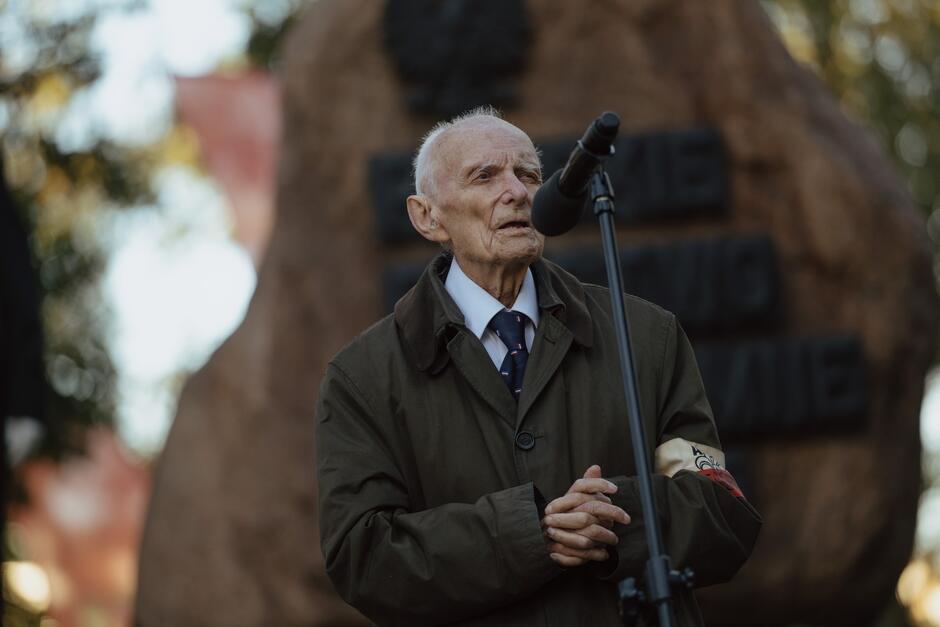 Na zdjęciu widać starszego mężczyznę przemawiającego do mikrofonu podczas oficjalnej uroczystości. Ma na sobie elegancki strój – ciemny płaszcz i krawat, a na ramieniu widoczna jest biało-czerwona opaska z symbolami, co sugeruje, że może to być uczestnik wydarzeń historycznych, prawdopodobnie były żołnierz Armii Krajowej lub innej formacji związanej z Polskim Państwem Podziemnym. Wyraz jego twarzy jest poważny, a cała scena ma bardzo podniosły charakter. W tle za mężczyzną znajduje się duży pomnik z napisem „Polskie Państwo Podziemne 1939-1945”, co dodatkowo wskazuje na to, że przemówienie związane jest z upamiętnieniem tego okresu w polskiej historii. Atmosfera zdjęcia jest pełna szacunku i nostalgii, a cała sytuacja ma wyraźny wymiar patriotyczny i historyczny.