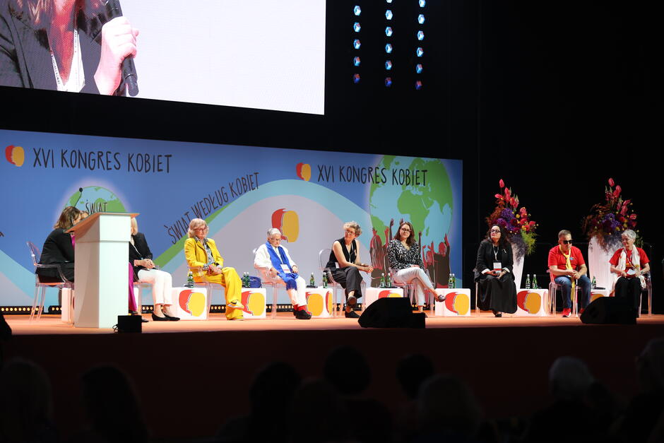 
Na zdjęciu widzimy panel dyskusyjny na scenie podczas XVI Kongresu Kobiet, gdzie siedem osób uczestniczy w rozmowie, a za nimi widnieje hasło „Świat według kobiet”. Uczestnicy siedzą na krzesłach, a scena jest udekorowana kolorowymi kwiatami, co podkreśla uroczysty i oficjalny charakter wydarzenia.