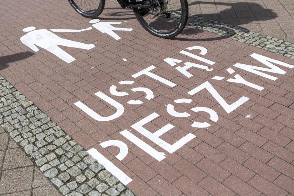 Na zdjęciu widoczna jest ścieżka rowerowa z namalowanym białym napisem USTĄP PIESZYM  oraz symbolami pieszych, sugerującymi pierwszeństwo dla pieszych. Częściowo widoczny jest również rower, który znajduje się nad napisem, co wskazuje, że zdjęcie zrobiono w miejscu wspólnego ruchu rowerowego i pieszego.