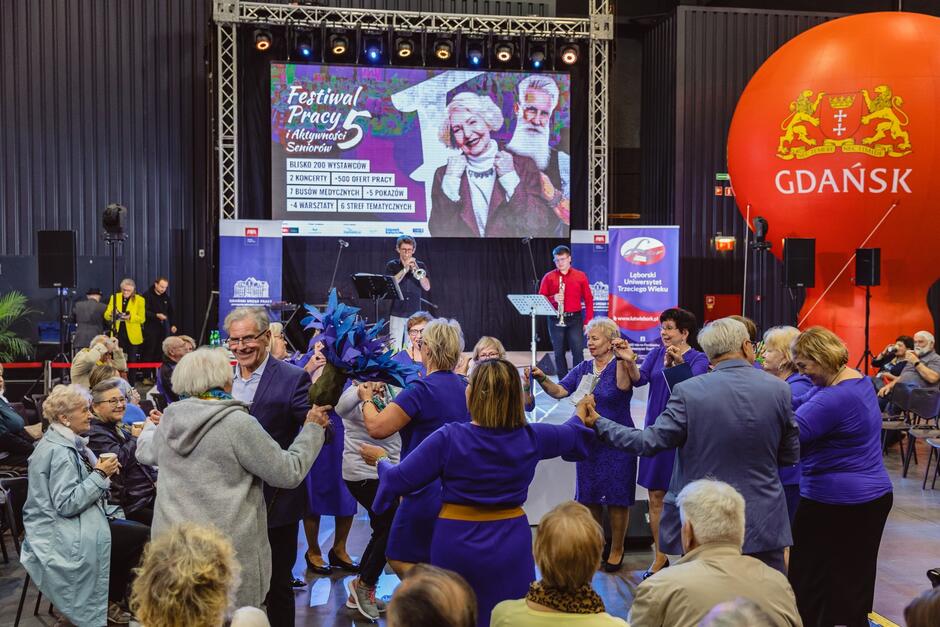 na zdjęciu grupa kilkunastu seniorów tańczy w kółeczku, w tle ogromny telebim