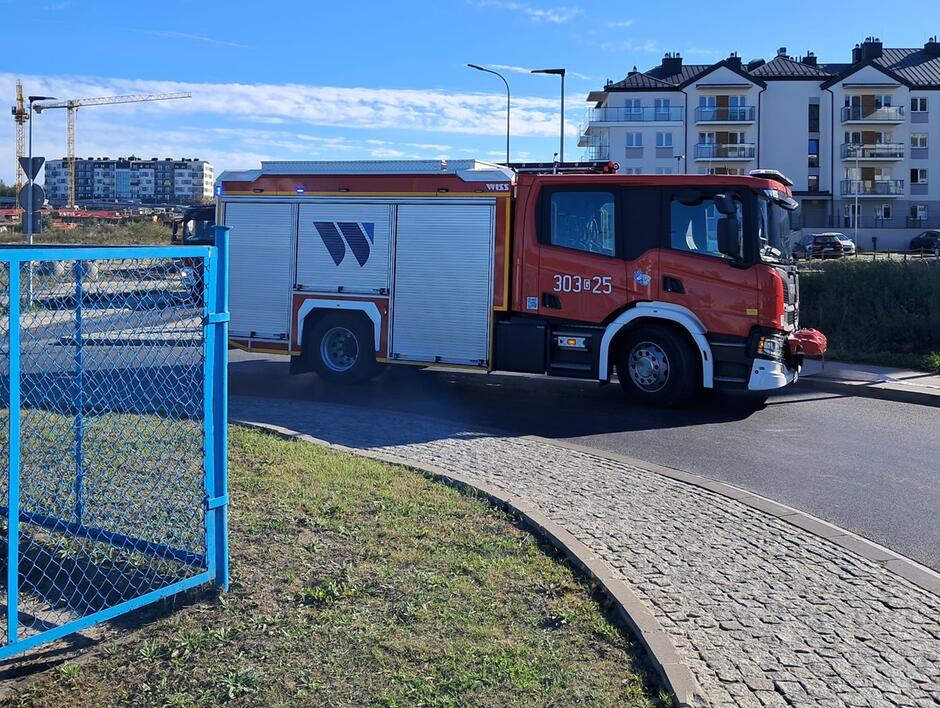 Na zdjęciu widać wóz strażacki zaparkowany na ulicy w okolicy osiedla mieszkalnego, na tle kilku bloków. Pojazd ma czerwony kolor, a na jego boku widoczny jest numer 303[25], co sugeruje przynależność do jednostki straży pożarnej w Polsce.