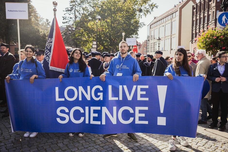 Na zdjęciu widoczna jest grupa młodych ludzi w niebieskich bluzach, trzymających duży baner z napisem LONG LIVE SCIENCE!  podczas akademickiej uroczystości. W tle widać osoby w togach oraz inne elementy wskazujące na to, że jest to wydarzenie związane z uczelniami, najprawdopodobniej Politechniką Gdańską, której nazwa widnieje na jednym z szyldów.