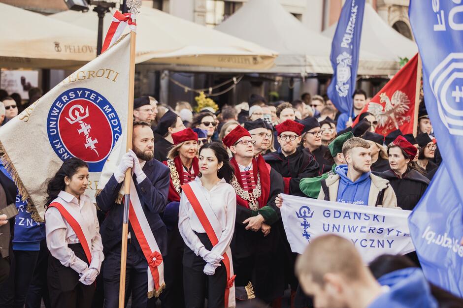 Na zdjęciu widoczna jest grupa osób uczestniczących w uroczystości akademickiej, najprawdopodobniej inauguracji roku akademickiego, związanej z Gdańskim Uniwersytetem Medycznym. Uczestnicy są ubrani w oficjalne stroje akademickie, w tym togi i birety, a niektórzy niosą sztandary uczelni.