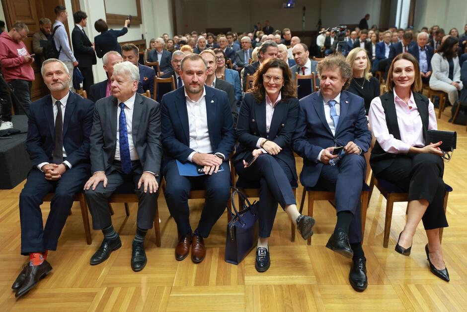 Zdjęcie przedstawia grupę elegancko ubranych osób siedzących w pierwszym rzędzie na wydarzeniu, najprawdopodobniej oficjalnej uroczystości akademickiej lub konferencji. Osoby na zdjęciu mają na sobie garnitury i formalne stroje. W tle widać więcej uczestników siedzących na krzesłach, a z tyłu po lewej stronie kilka osób fotografuje wydarzenie. Atmosfera wydaje się formalna, a uśmiechy na twarzach niektórych osób sugerują pozytywną lub podniosłą okazję. Pomieszczenie ma drewnianą podłogę i wydaje się być dużą salą, prawdopodobnie konferencyjną lub audytoryjną.