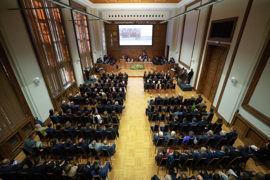 Zdjęcie przedstawia dużą salę akademicką, w której odbywa się oficjalna uroczystość, prawdopodobnie inauguracja roku akademickiego. Widok został uchwycony z góry, co pozwala zobaczyć całą przestrzeń. Na środku sali ustawione są rzędy krzeseł, na których siedzą uczestnicy wydarzenia. W pierwszych rzędach znajdują się osoby w formalnych strojach, a z przodu, na podwyższeniu, widzimy stół prezydialny, przy którym siedzą przedstawiciele uczelni w togach i insygniach akademickich. Na ścianie za nimi znajduje się duży ekran, na którym wyświetlono slajd z informacjami dotyczącymi wydarzenia. Cała sala jest elegancka, z wysokimi oknami i drewnianym wykończeniem, co nadaje uroczysty charakter tej ceremonii.