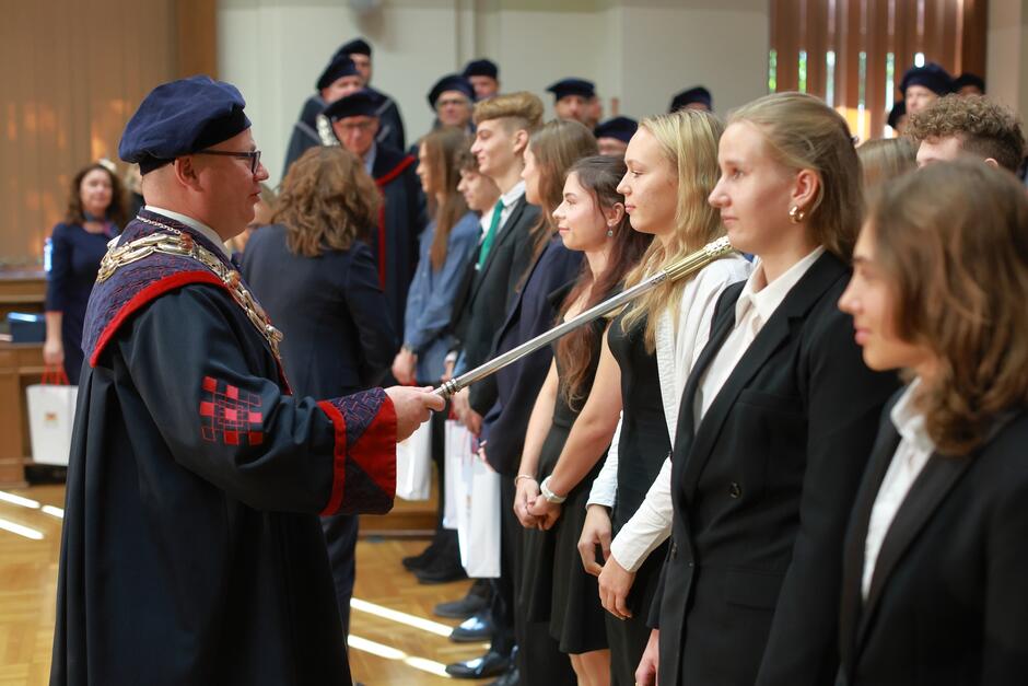  Zdjęcie przedstawia moment symbolicznego pasowania, najprawdopodobniej podczas ceremonii immatrykulacji studentów. Osoba w tradycyjnym stroju akademickim, prawdopodobnie rektor lub inny przedstawiciel uczelni, trzyma ceremonialny miecz, którym dotyka ramienia jednej ze stojących w rzędzie młodych osób, ubraną w formalny strój. To rytuał mający na celu włączenie nowych studentów w poczet uczelni. W tle widać innych członków władz uczelni, również ubranych w togi i birety. Cała scena ma uroczysty charakter i odbywa się w eleganckiej sali, co podkreśla powagę i znaczenie ceremonii.